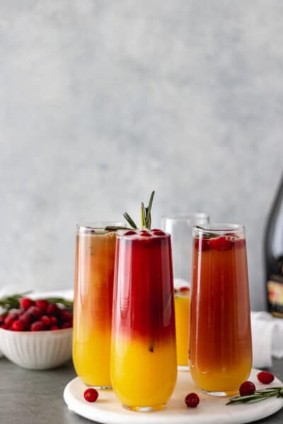 Three Christmas Cranberry Mimosas garnished with fresh cranberries and a rosemary sprig, which is surrounded by a prosecco bottle and a bowl of cranberries.