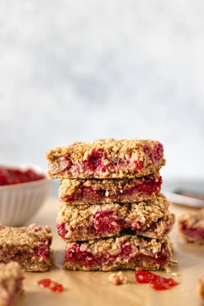 A stack of four strawberry crumble bars on parchment paper surrounded by other crumble bars and a pinch bowl of strawberry spread.