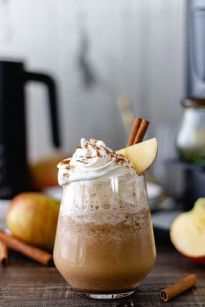 Apple Pie Espresso Frappuccino, in a glass, surrounded by apples, cinnamon sticks and a coffee machine.