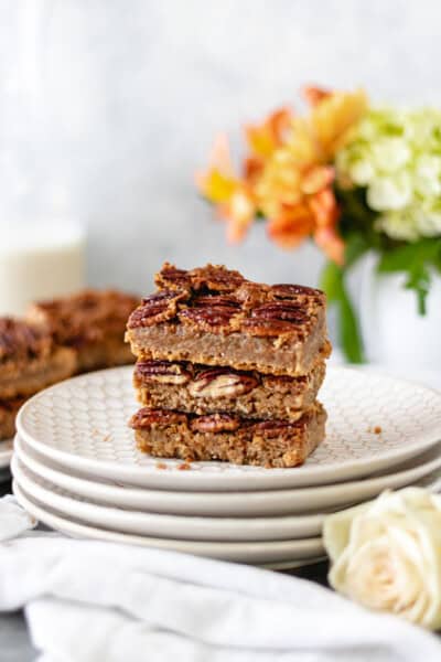 A stack of pecan pie bars on a stack of plates surrounded by fresh flowers and milk.