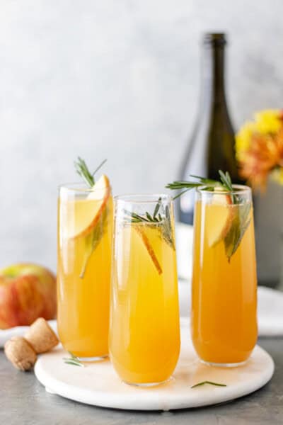 Three Apple Cider Mimosas garnished with an apple slice and a rosemary sprig, which is surrounded by a prosecco bottle and flowers.