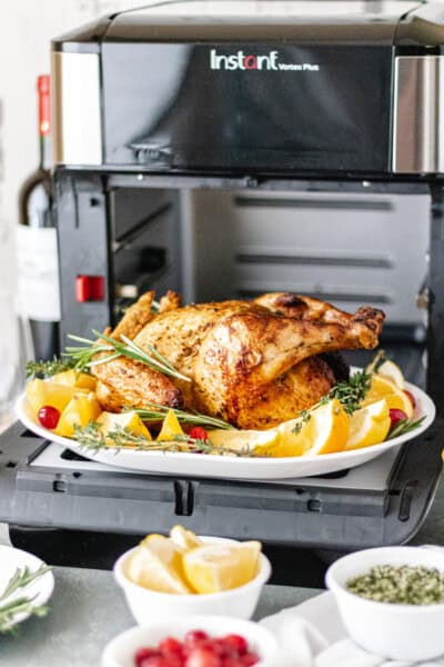 Whole roasted chicken on a platter, sitting on the open door of an air fryer oven alongside pinch bowls filled with lemon slices, fresh cranberries, and herb salt.