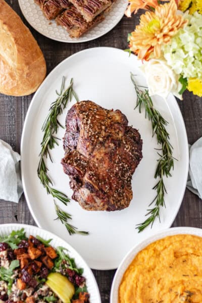 Herb-Roasted Boneless Leg of Lamb on a platter surrounded by salad, mashed sweet potatoes, pecan pie bars and fresh flowers.
