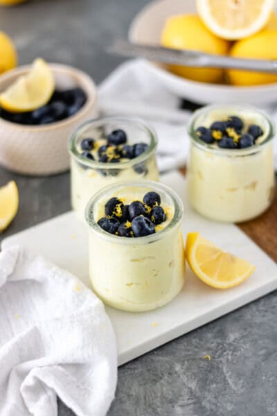 Three mini glass jars of lemon curd topped with fresh blueberries and lemon zest and sitting on a small marble board.