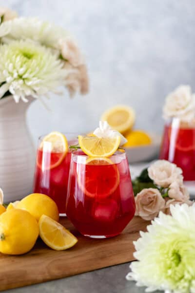 Iced Passion Tea Lemonade in glasses sitting on a cutting board and surrounded by fresh flowers and lemons.