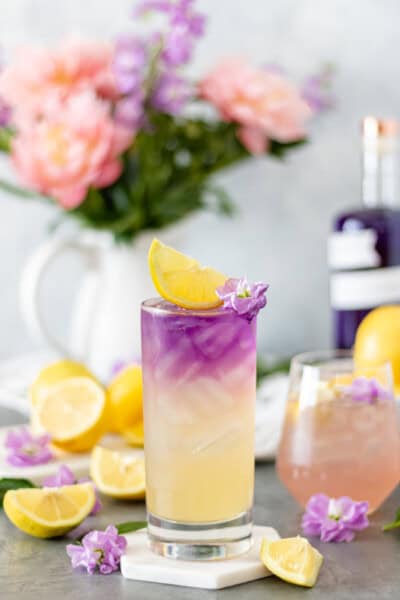 Two glasses of Empress Gin Lemonade surrounded by fresh lemons, flowers and a bottle of Empress 1908 Gin.
