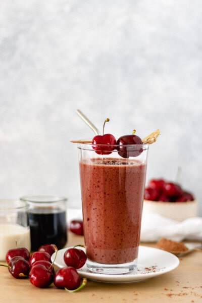 Chocolate-Cherry Cold Brew Smoothie in a glass topped with cacao nibs and fresh cherries, that is sitting on a mini plate, which is surrounded by fresh cherries, cacao powder, and mini glasses of milk and cold brew.