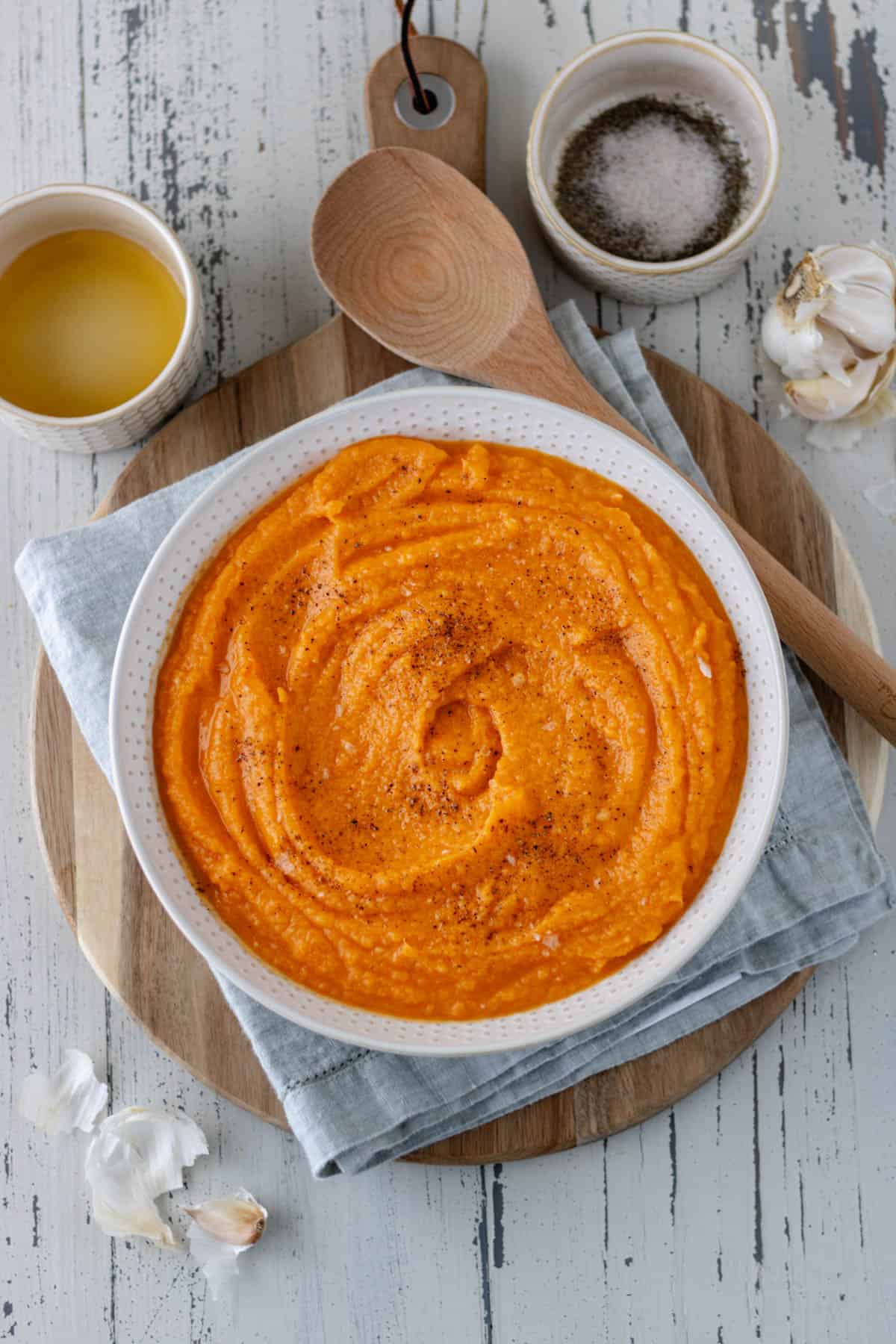 Delicious Mashed Sweet Potatoes in a large serving bowl, topped with black pepper and salt, and sitting on a folded towel and circular wood bread board.