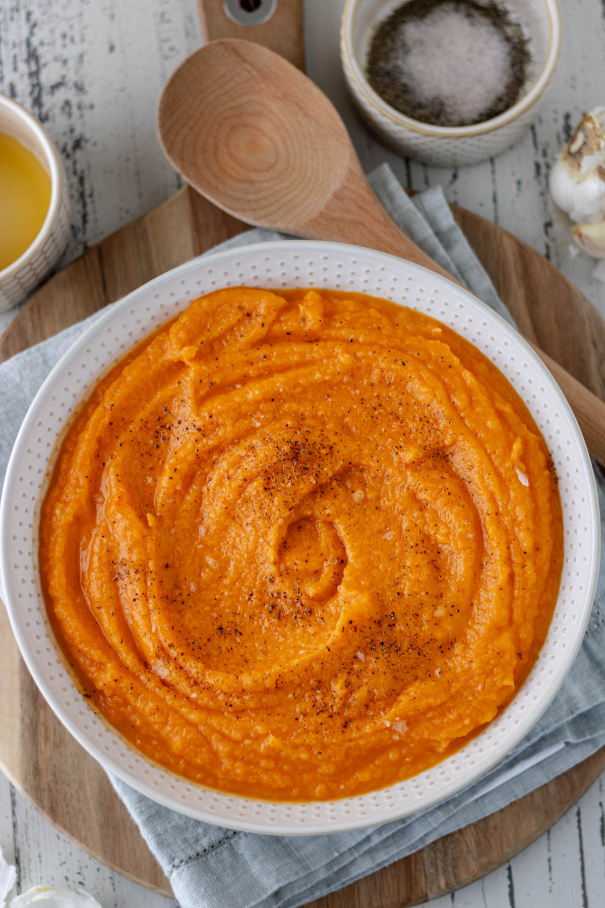 A large serving bowl of mashed sweet potatoes, sitting on a circular wood bread board and surrounded by a head of garlic and a ramekin of maple syrup and a ramekin of salt and pepper.