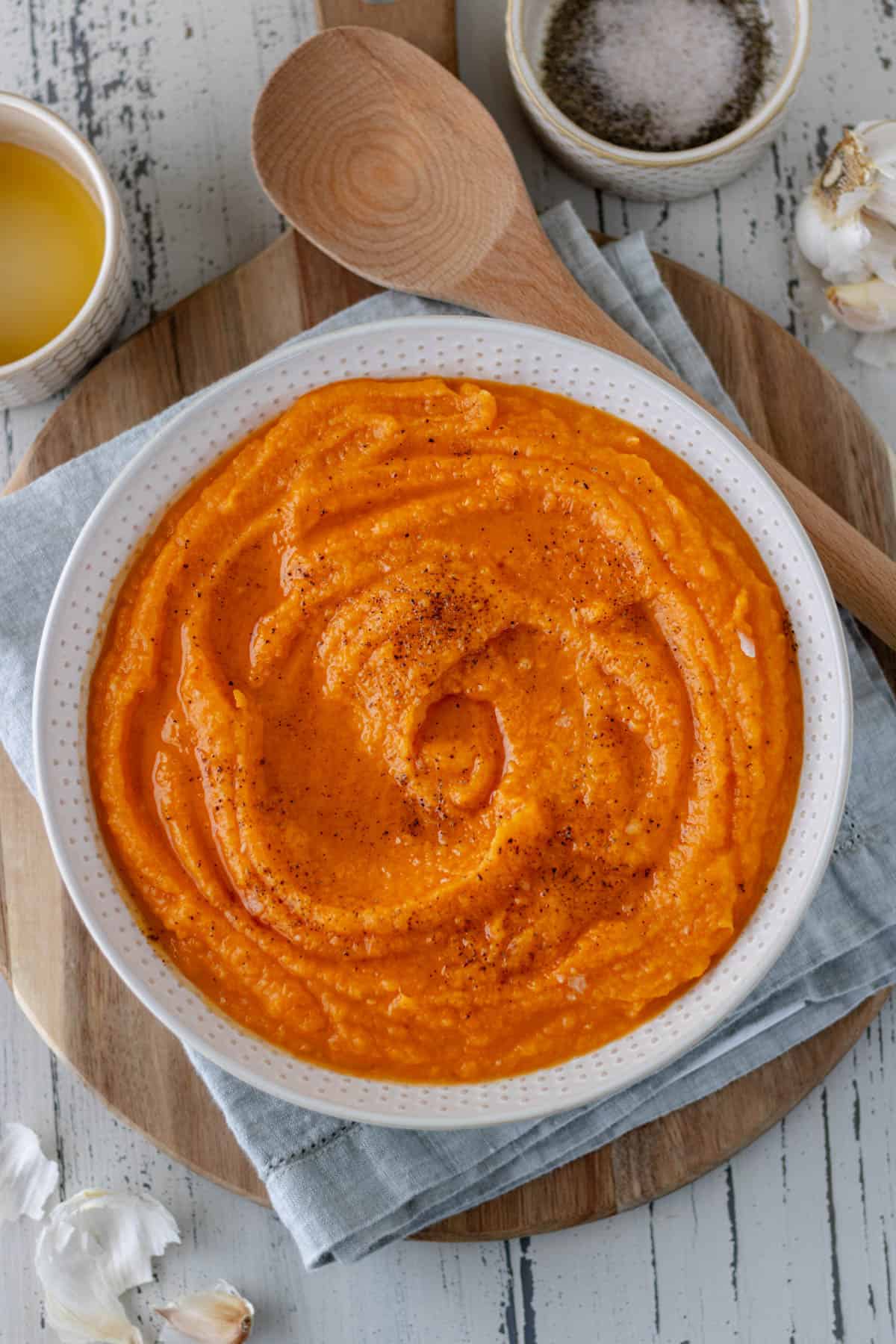 Delicious Mashed Sweet Potatoes in a large serving bowl, topped with black pepper, salt and maple syrup, and sitting on a folded towel and circular wood bread board.