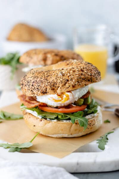 A BLT Fried Egg Bagelwich with a slightly runny yolk alongside a glass of orange juice and a bowl of arugula.