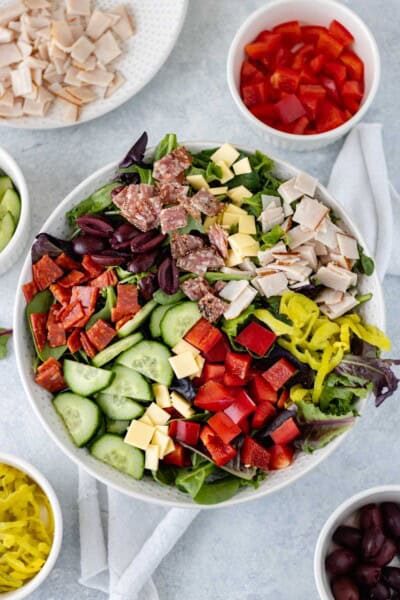 Mixed greens in a large serving bowl topped with diced meats, cucumber, bell pepper, pepperoncini, dairy-free cheese and olives and surrounded by individual bowls of the same ingredients.