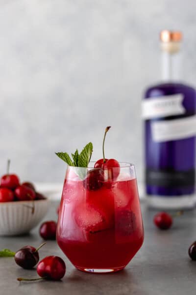 A glass of a Cherry-Berry Gin Kombucha Cocktail surrounded by fresh cherries and a bottle of Empress 1908 Gin.