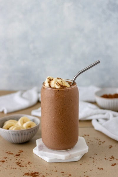 A glass of Chocolate-Peanut Butter Breakfast Smoothie topped with sliced bananas and sprinkled with cacao powder and sitting on a stack of marble coasters.