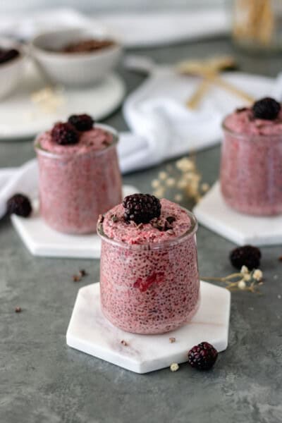 Three mini glass jars of Roasted Strawberry Chia Pudding, sitting on marble coasters, alongside pinch bowls of cacao nibs and blackberries.