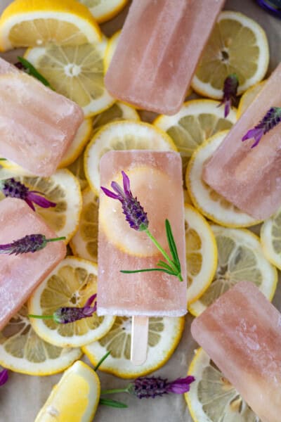 Six Lavender Gin Lemonade Popsicles in a circle, topped with fresh lavender flowers, and laying on a bed of fresh lemon slices.