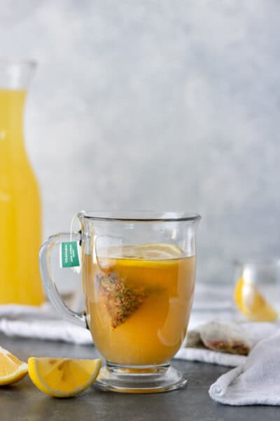 A full glass mug of Hot Honey Ginger Tea surrounded by lemon slices, tea bags and a large pitcher of homemade lemonade.