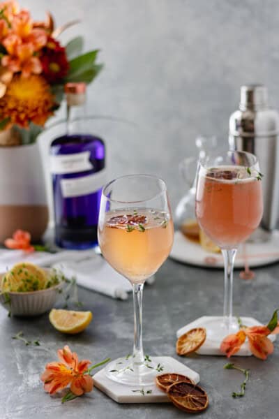 Two champagne glasses filled with gin, simple syrup, lemon juice and prosecco, and surrounded by fresh flowers, lemon slices and thyme.