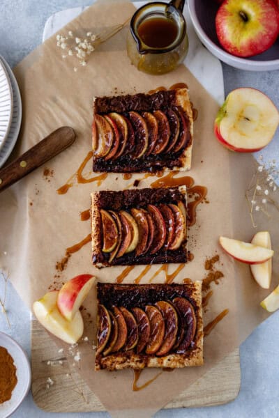 Three mini Honeycrisp-Apple Tarts, in a row, drizzled with caramel sauce and surrounded by apple slices, a mini glass pot of caramel sauce and baby's breath.