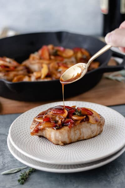A cooked pork chop with apple and plum slices, sitting on a plate and being drizzled with sauce.