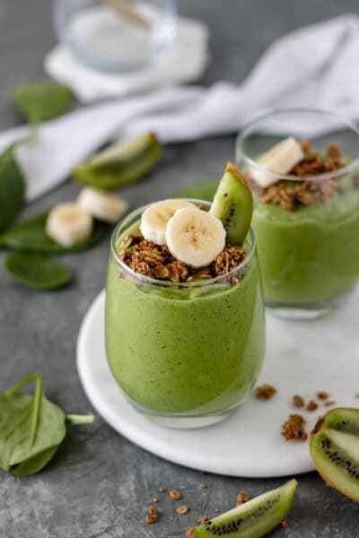 Two glasses of a Kiwi Avocado Green Smoothie topped with granola, a kiwi wedge and banana slices.