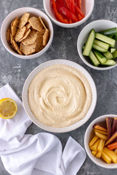 A bowl of homemade hummus surrounded by smaller bowls of crackers, bell pepper slices, cucumber slices and carrot slices.