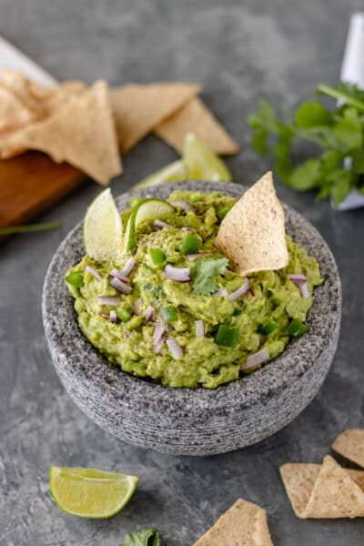 Avocado Serrano Guacamole in a mortar and topped with diced red onion, diced serrano pepper, a lime slice, and a tortilla chip.