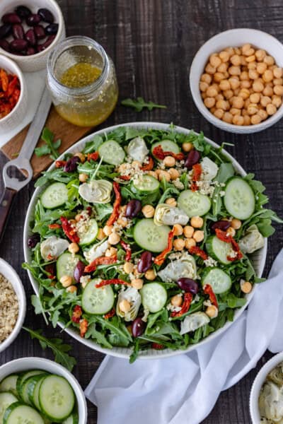 Mediterranean Quinoa Salad in a large bowl and surrounded by small pinch bowls of sundried tomatoes, quinoa, cucumber slices, artichoke hearts, garbanzo beans, a homemade vinaigrette and Kalamata olives.