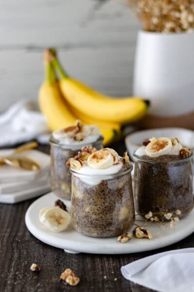 Three mini glass jars filled with Banana-Nut Bread Chia Pudding, sitting on a marble trivet, and surrounded by a vase of baby’s breath, a bunch of bananas, fresh banana slices and walnuts.