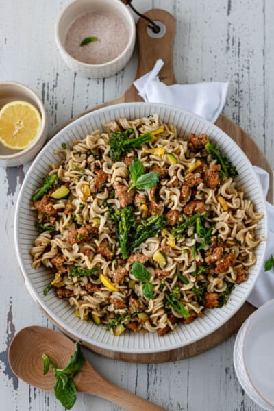 Sausage Squash and Broccolini Pasta in a large ceramic bowl, sitting on top of a circular wood board, and surrounded by a wooden spoon and two pinch bowls of salt and half a lemon.