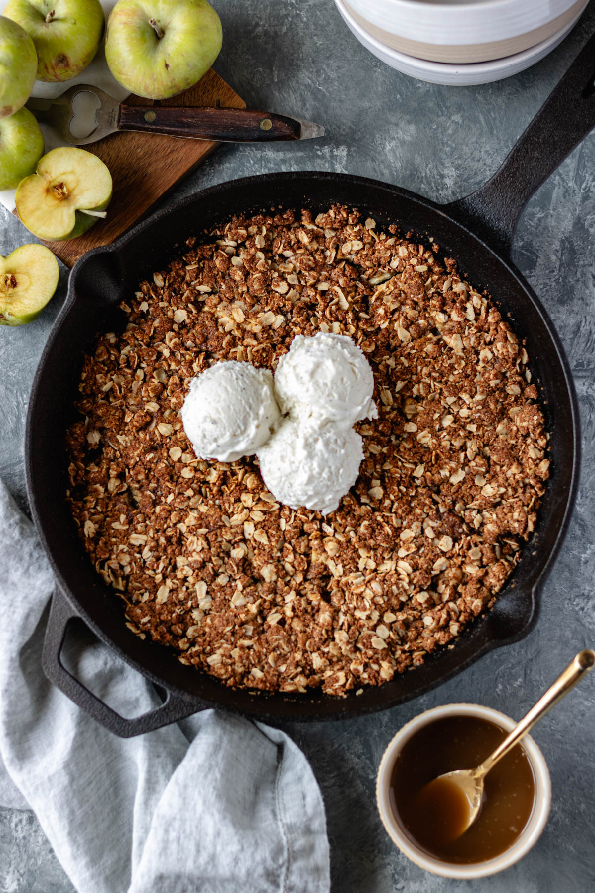 Baked apples topped with an oatmeal mixture, in a cast iron skillet, surrounded by a pinch bowl of vegan caramel sauce, a stack of bowls, a cutting board with green apples and a cloth napkin.