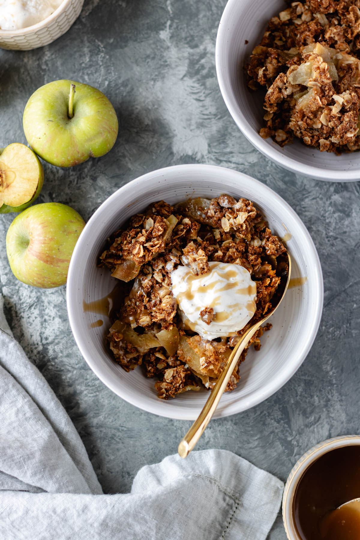 Gluten-Free Apple Crisp in a bowl topped with dairy-free vanilla ice cream and caramel sauce and surrounded by apples, a pinch bowl of ice cream, a pinch bowl of vegan caramel sauce and a second bowl of apple crisp.