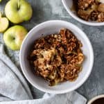 Baked apples topped with an oatmeal mixture, in a bowl, surrounded by apples, a pinch bowl of dairy-free vanilla ice cream, a pinch bowl of vegan caramel sauce, a second bowl of apple crisp and a cloth napkin.