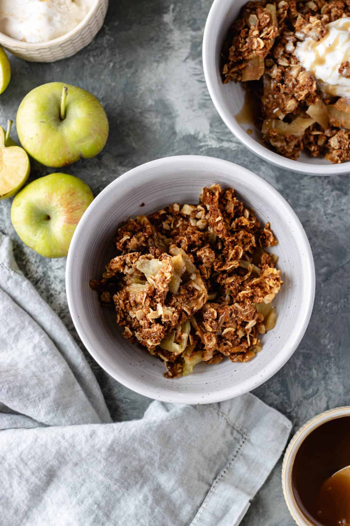 Baked apples topped with an oatmeal mixture, in a bowl, surrounded by apples, a pinch bowl of dairy-free vanilla ice cream, a pinch bowl of vegan caramel sauce, a second bowl of apple crisp and a cloth napkin.