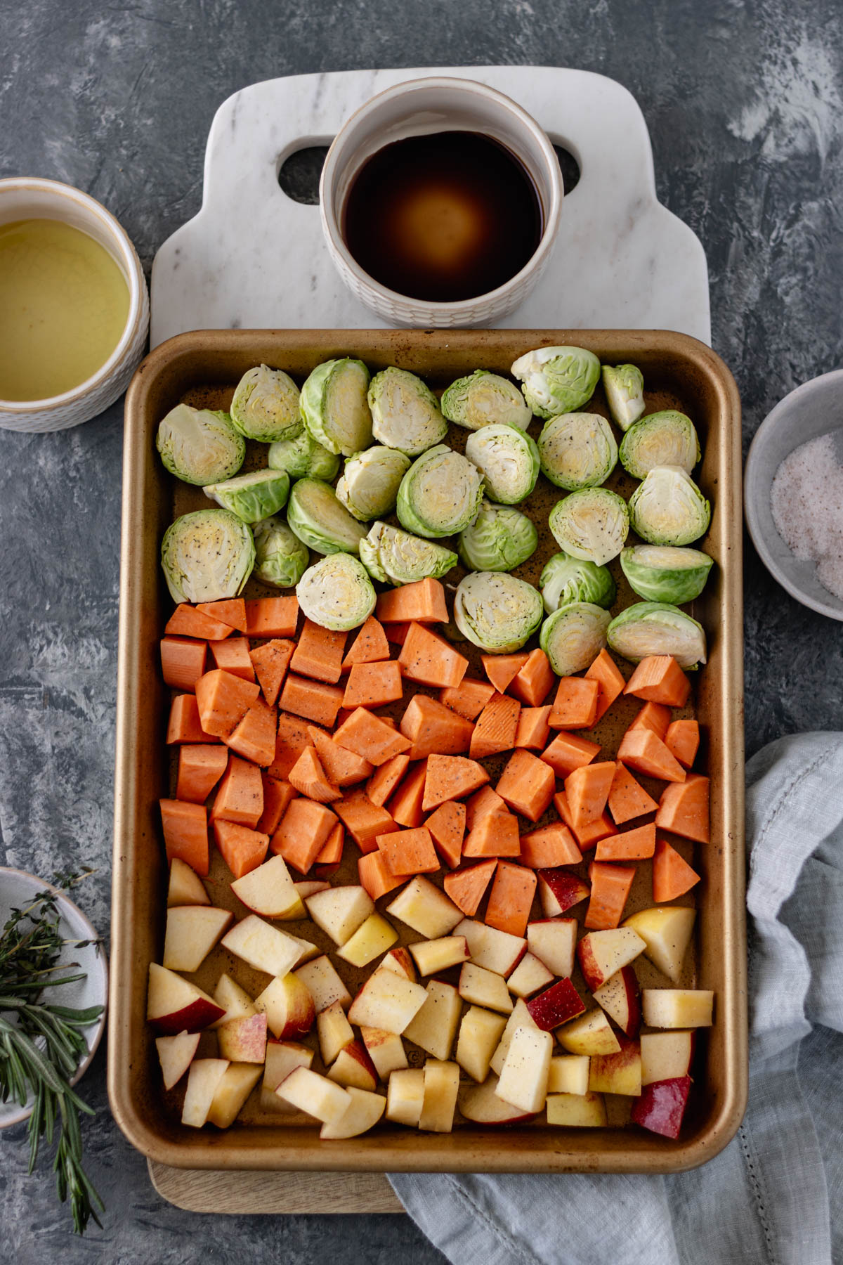 A sheet pan of diced brussels sprouts, sweet potatoes and apples, sitting on a marble bread board, and surrounded by pinch bowls of salt, balsamic, extra virgin olive oil and herbs.