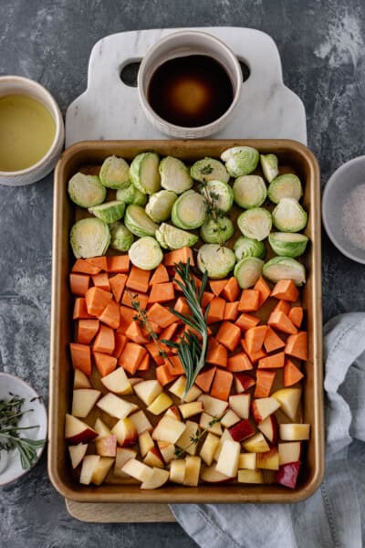 A sheet pan of diced brussels sprouts, sweet potatoes and apples, topped with fresh rosemary and thyme sprigs, and surrounded by pinch bowls of salt, balsamic, extra virgin olive oil and herbs.