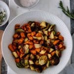 Cooked Maple-Herb-Roasted Sheet Pan Vegetables in a large ceramic bowl, sitting on a marble bread board, and surrounded by pinch bowls of salt, herbs and a hand towel.