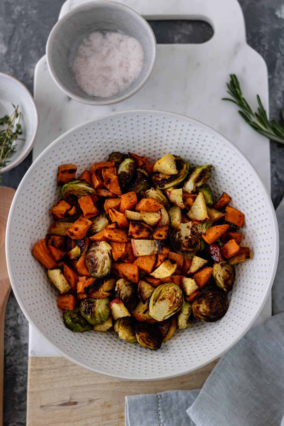Cooked Maple-Herb-Roasted Sheet Pan Vegetables in a large ceramic bowl, sitting on a marble bread board, and surrounded by pinch bowls of salt, herbs and a hand towel.
