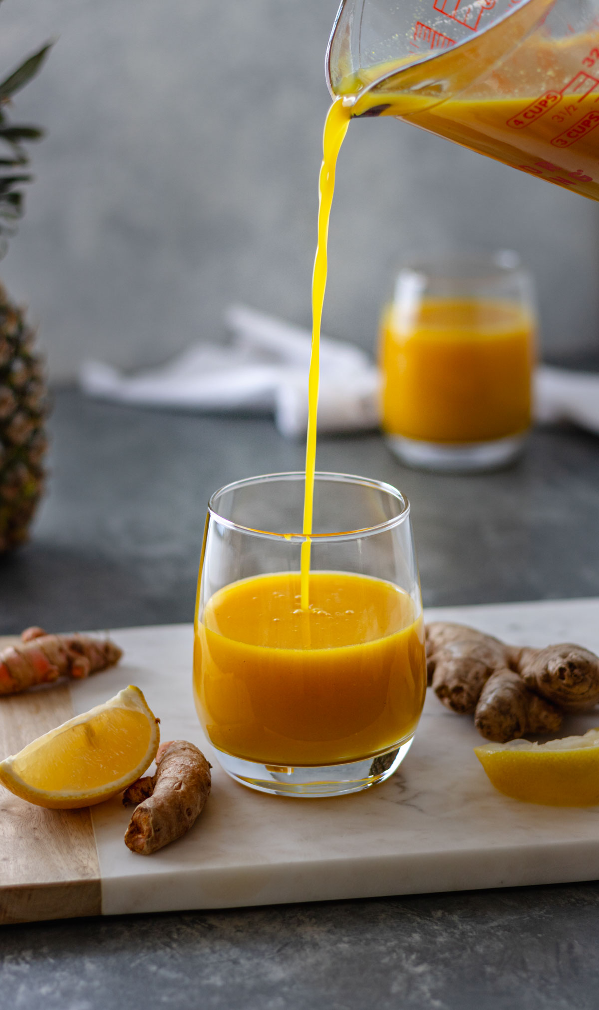 A 4-cup glass measuring cup pouring pineapple, lemon juice into a glass, surrounded by ginger root, turmeric root, and lemon wedges that are all sitting on a rectangular marble bread board.