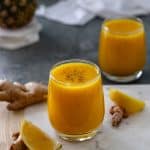 An Immune Boosting Wellness Shot in a glass, topped with black pepper, surrounded by lemon wedges, turmeric, and ginger root, alongside another glass of juice, a tea towel and a whole pineapple sitting on a stack of marble coasters.