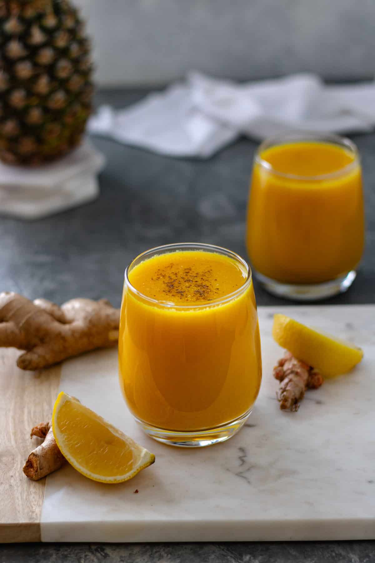 An Immune Boosting Wellness Shot in a glass, topped with black pepper, surrounded by lemon wedges, turmeric, and ginger root, alongside another glass of juice, a tea towel and a whole pineapple sitting on a stack of marble coasters.