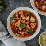 Three glass bowls filled with broth, cooked chicken, diced sweet potato, black beans, and roasted tomatoes, surrounded by a cloth napkin, a bunch of cilantro and a pinch bowl of lime wedges.