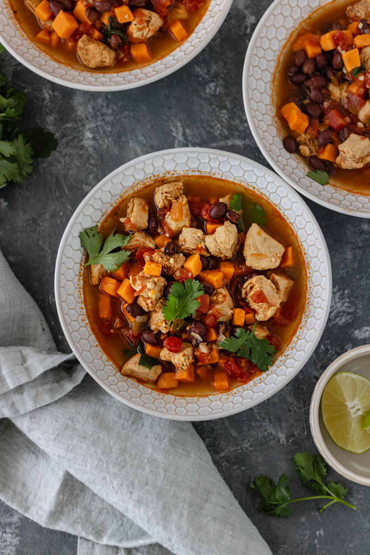 Three glass bowls filled with broth, cooked chicken, diced sweet potato, black beans, and roasted tomatoes, surrounded by a cloth napkin, a bunch of cilantro and a pinch bowl of lime wedges.
