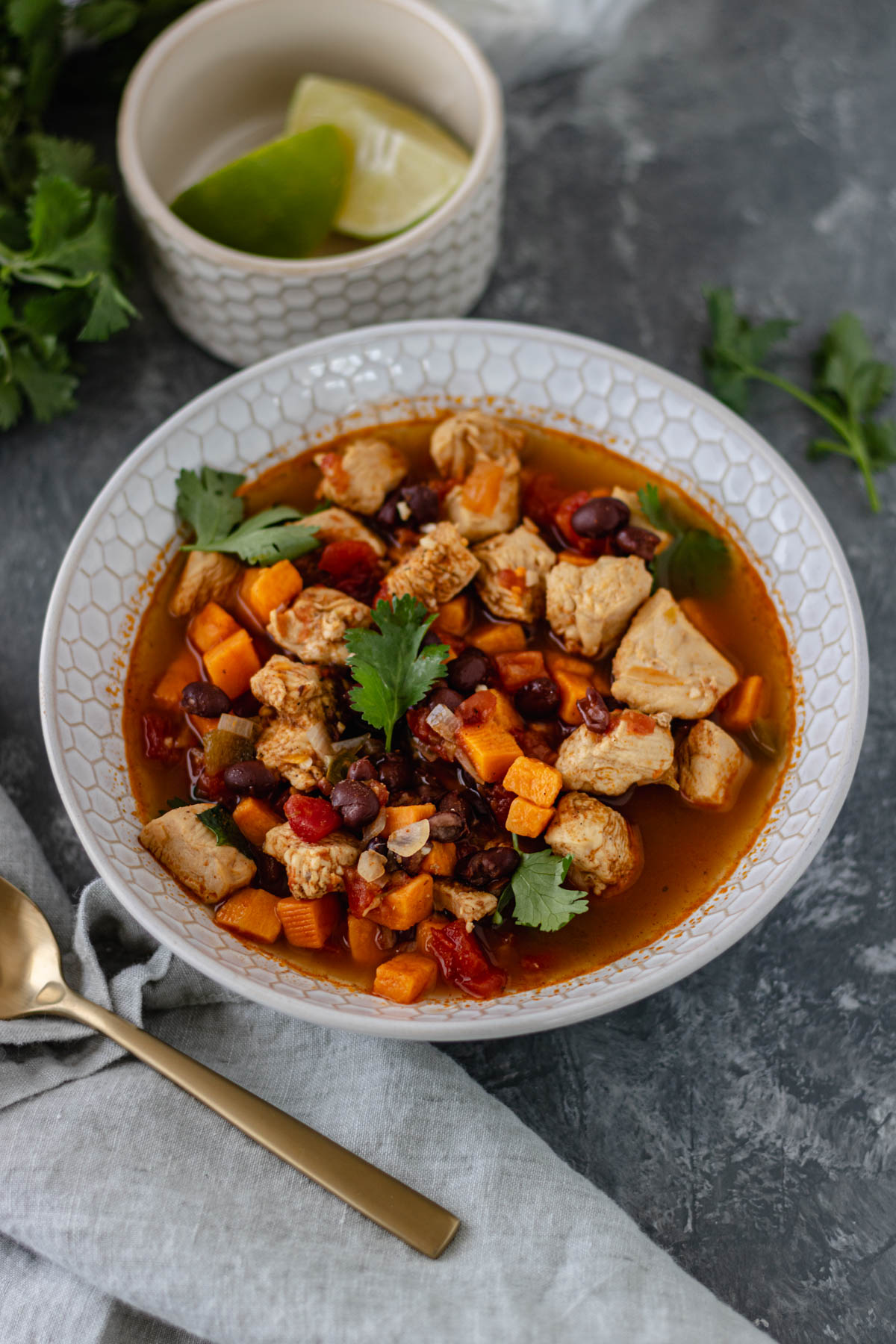 A glass bowl filled with broth, cooked chicken, diced sweet potato, black beans, and roasted tomatoes, topped with cilantro, and surrounded by a cloth napkin, spoon, and more cilantro.