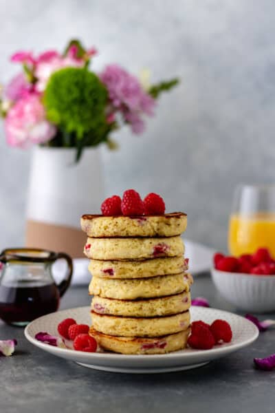 A stack of seven, Fluffy Raspberry Almond Pancakes, on a salad plate, surrounded by a pinch bowl of fresh raspberries, glass of orange juice, a vase of pink and green flowers and a mini glass pot filled with maple syrup.