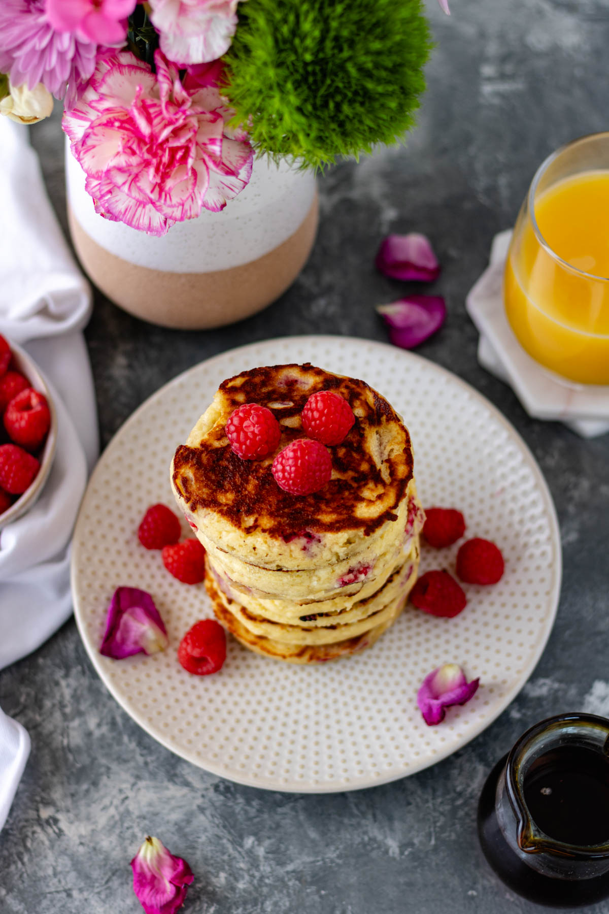 A stack of pancakes on a salad plate topped with fresh raspberries and surrounded by more raspberries and pink flower petals.