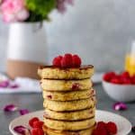 A stack of pancakes drizzled with maple syrup, topped with fresh raspberries, and surrounded by more raspberries and pink flower petals.