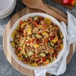 One Skillet-Sausage and Veggies with Cauliflower Rice in a large serving bowl that is sitting on a circular bread board, and surrounded by a white tea towel, serving spoon, ceramic bowls and a mini cutting board with mini bell peppers and sliced zucchini on top.