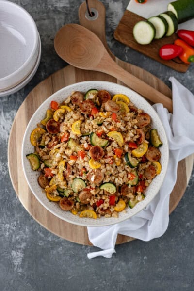 One Skillet-Sausage and Veggies with Cauliflower Rice in a large serving bowl that is sitting on a circular bread board, and surrounded by a white tea towel, serving spoon, ceramic bowls and a mini cutting board with mini bell peppers and sliced zucchini on top.