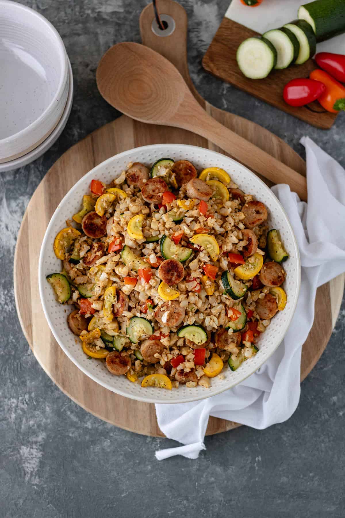 One Skillet-Sausage and Veggies with Cauliflower Rice in a large serving bowl that is sitting on a circular bread board, and surrounded by a white tea towel, serving spoon, ceramic bowls and a mini cutting board with mini bell peppers and sliced zucchini on top.